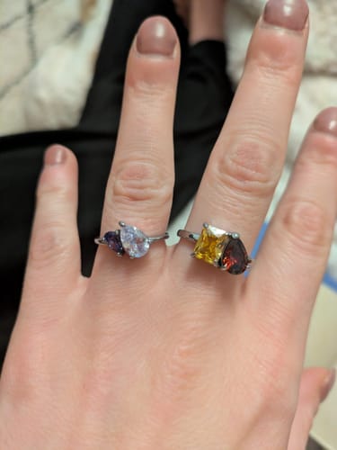 Customer's hand wearing the Leesha Birthstone Ring with purple and clear stones on their index finger, next to another ring.