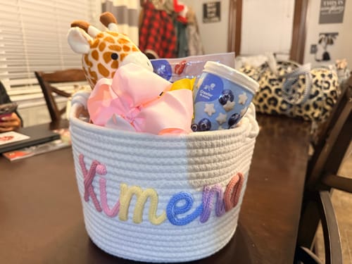 Customer's personalized white rope basket with the name 'Ximena' in colorful yarn, filled with baby gifts on a table.