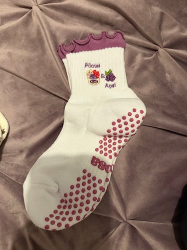 Reviewer's white Pilates & Açaí socks with a purple ruffled cuff and grip dots, laid out on a purple tufted background.