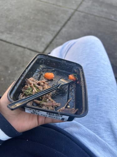 Customer holding a partially eaten Beef Pot Roast meal in its black container, with a fork resting inside.