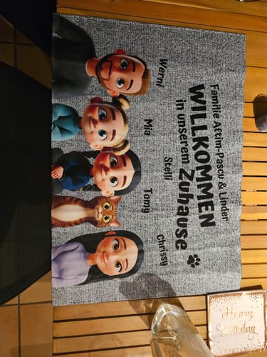 Kundenfoto der personalisierten 'Unser Zuhause'-Fußmatte mit Cartoon-Figuren einer Familie auf einem Holzboden.