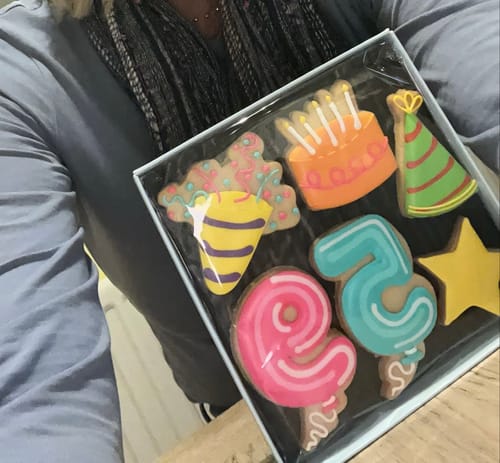 Reviewer holding the Birthday Balloon Biscuit Box with colorful iced biscuits shaped like a cake, a hat, and the numbers 6 and 5.