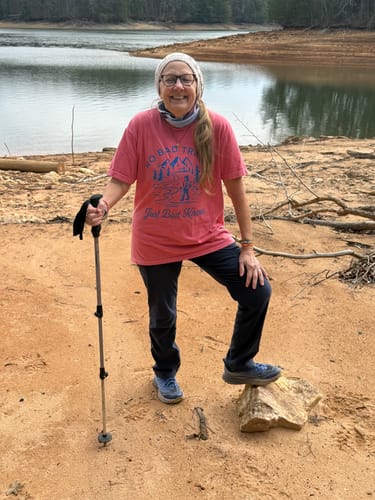 Customer wearing the pink Bad Knees T-shirt, posing with a hiking pole by a lake.