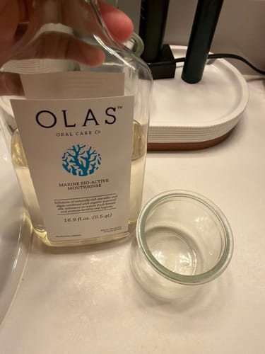 Customer's hand holding a clear glass bottle of OLAS Mouthrinse next to an empty glass cup on a counter.
