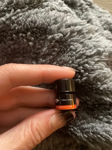 Reviewer's hand holding a tiny sample bottle of Troll Food perfume against a soft grey textile.