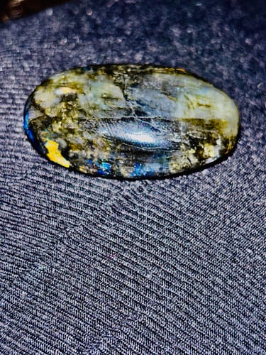 Close-up: Customer's iridescent oval crystal with blue and yellow hues on a dark fabric surface.