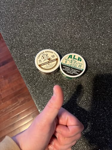 Reviewer gives a thumbs-up to tins of Mountain Wintergreen Nicotine Pouches and Coffee Break pouches on a counter.