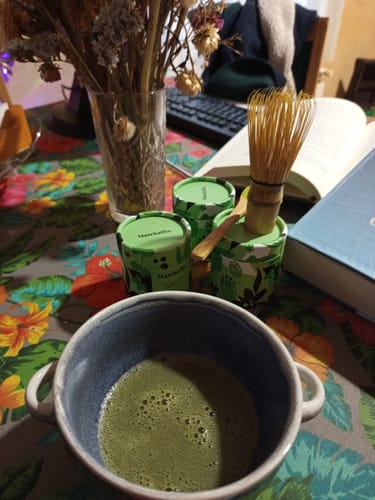 Cliente muestra su Té Matcha Premium preparado en una taza, con los envases y un batidor de bambú sobre una mesa.