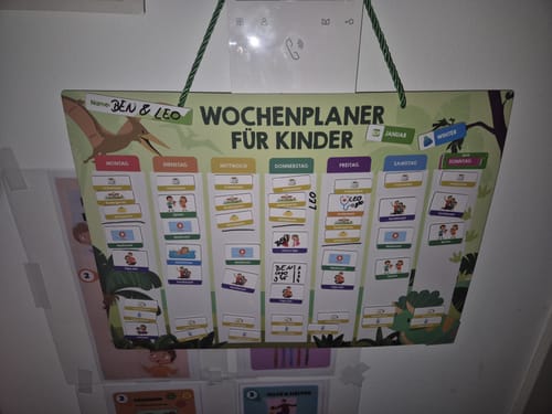 Ein Rezensent zeigt den bunten magnetischen Wochenplaner für Kinder, der mit Aktivitäten gefüllt an der Wand hängt.