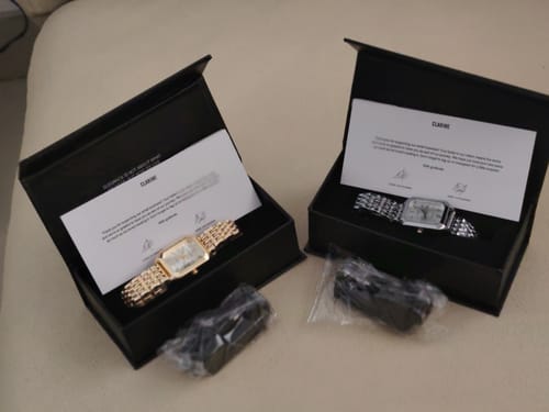 Reviewer's two CLAIRE DE MAINS watches, one gold and one silver, displayed in their open black boxes.