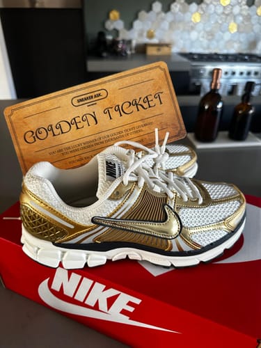 Recensent toont de Nike Zoom Vomero 5 Metallic Gold sneaker op de open schoenendoos met een gouden ticket erachter.