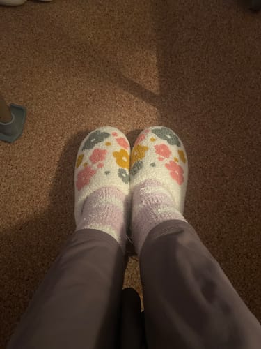 Reviewer's view of their feet wearing the Livia Flower Slippers with a colorful floral pattern on a brown carpet.