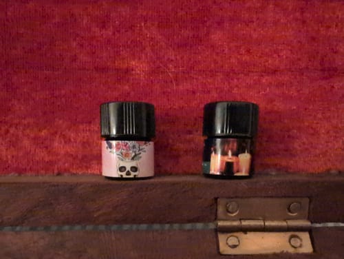 Reviewer's All's Fair perfume bottle with a skull and flower label, shown next to another bottle.