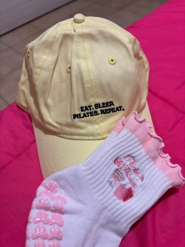 Reviewer's yellow 'Eat. Sleep. Pilates. Repeat.' cap lying on a bright pink surface next to a Pilates sock.