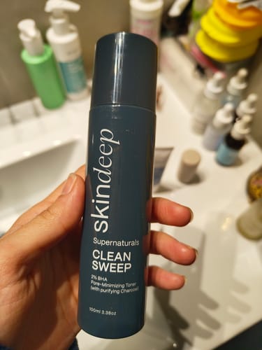 Reviewer holding the dark grey bottle of CLEAN SWEEP Pore-Minimizing Toner in a bathroom.