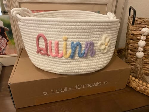 Customer's personalized white knitted basket with 'Quinn' in multicolor yarn and a flower icon.