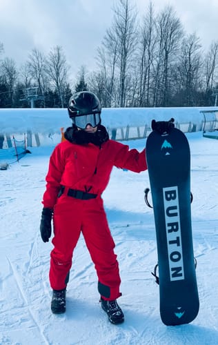Reviewer wearing the red one-piece ski suit while holding a snowboard on a snowy slope.