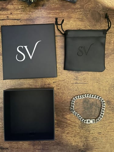 Un cliente muestra la pulsera Cuban de 8mm de acero sobre una mesa de madera junto a su caja y funda negras de la marca.