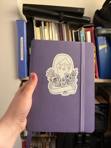 Customer holding their periwinkle Weekly 2026 Planner, which has a large illustrated sticker on the cover.