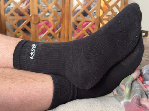 Customer relaxing with feet crossed, wearing the black crew socks which show a snug fit and the embroidered logo.