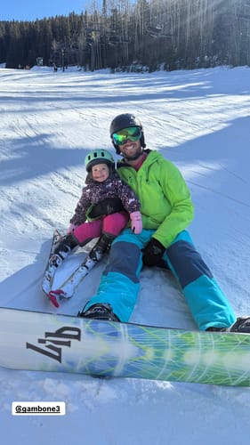 Customer sitting on a snowy slope wearing the color-block Waterproof Snow Bibs with a snowboard.