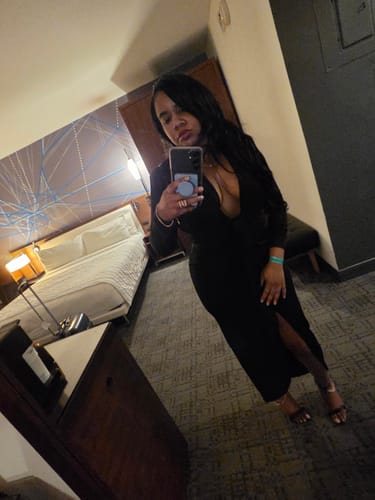 Reviewer in a hotel room wearing the long black Brielle dress, showing the deep plunge neckline and front slit.