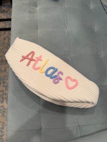 Customer's personalized knitted basket shown collapsed on a couch, with the name 'Atlas' and a heart.