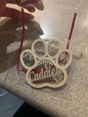 Customer holding a personalized wooden Christmas ornament shaped like a paw print with the name "Cuddles" in its package.