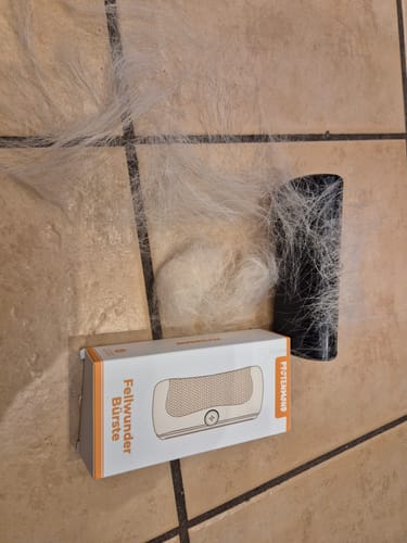 Ein Rezensent zeigt die Verpackung der Fellwunder Bürste neben einem großen Haufen ausgekämmten weißen Fells auf einem Fliesenboden.