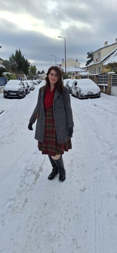 Une cliente porte la robe à carreaux rouge vin sous un manteau gris dans une rue enneigée.
