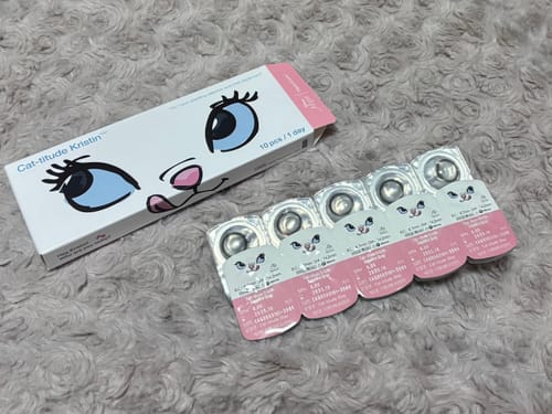 レビューアーによる、「Cat-titude Kristin 1Day」の箱とブリスターパック。ふわふわしたグレーの布の上に置かれている。