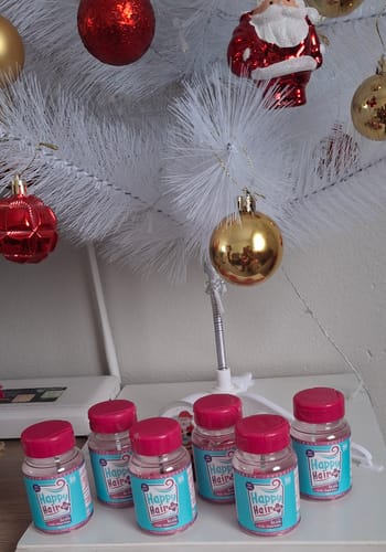 Cliente exibe sete potes do suplemento Happy Hair em frente a uma árvore de Natal branca.