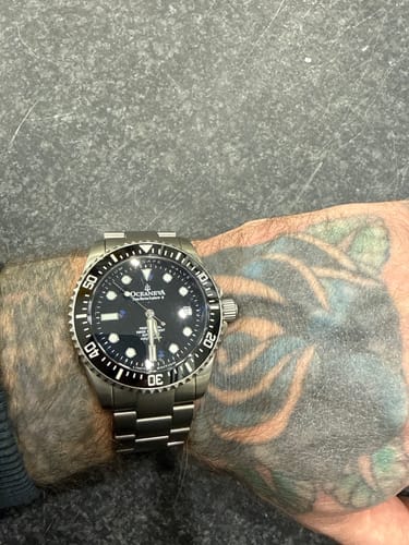 A customer's Oceaneva Deep Marine Explorer II titanium watch on their tattooed wrist, showing the black dial and bezel.