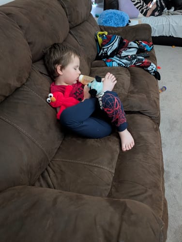 Reviewer's child relaxing on a brown couch, drinking from a bottle next to a red toy.