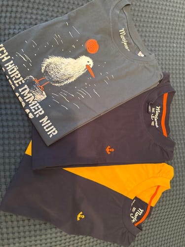 Ein Kunde zeigt drei gefaltete T-Shirts, darunter das graue 'Ich höre immer nur Mimimi' Bio-T-Shirt auf einem blauen Laken.