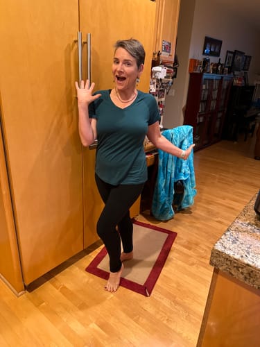 Reviewer wearing the green Mia V Neck shirt and black leggings while posing happily in a kitchen.
