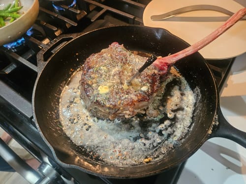 Reviewer basting a USDA Choice Tomahawk Ribeye with butter and herbs in a hot cast-iron skillet.