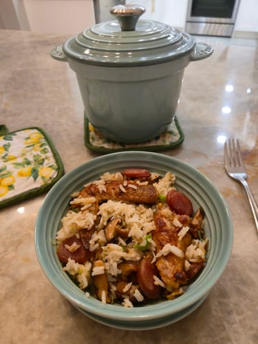 Reviewer's sage green Le Creuset Cast Iron Rice Pot on a countertop, with a prepared rice dish in a bowl in the foreground.