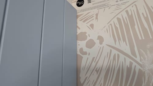 Close-up of a reviewer's The Palms Wallpaper in Ecru sample next to a blue paneled wall.