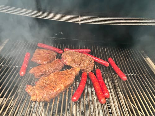 Customer's Aggie Grilling Bundle steaks and hotdogs smoking on a large grill.