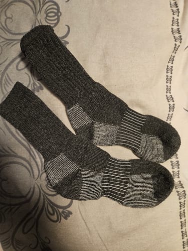 Rezensent zeigt ein Paar dicke, dunkelgraue Naturwide Merino Wolle Socken, die flach auf einem gemusterten Stoff liegen.