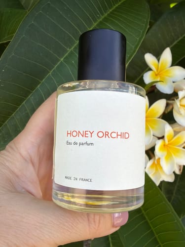 Customer holding a bottle of Honey Orchid perfume with a background of green leaves and white flowers.