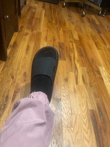Customer wearing one black, thick-soled Cloud Slide indoors on a wood floor.