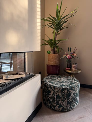 Klantfoto van de Woood Sara Botanical poef in een stijlvolle hoek naast een plant en een moderne haard.