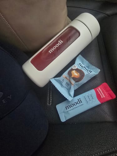 Customer's sachet of Enhanced Electrolytes next to a Moodi water bottle and snack on a black leather car seat.