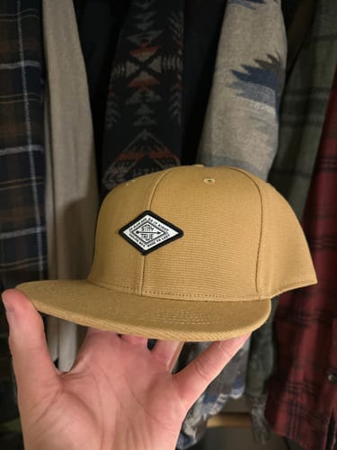 Customer holding the tan Big Flat Bill Structured Canvas Hat in their hand in front of clothes in a closet.