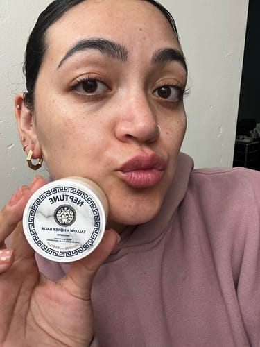 Customer holding a small jar of the Grass Fed Tallow & Honey Balm in front of her clear skin.