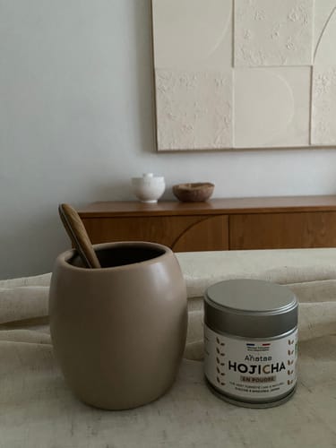 Une cliente montre sa boîte de THÉ HOJICHA BIO à côté d'une tasse beige avec une cuillère en bois.