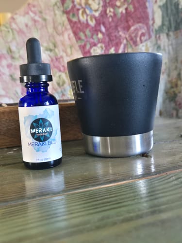 Reviewer's blue bottle of Meraki Blu Methylene Blue with its dropper, next to a black cup on a wooden table.