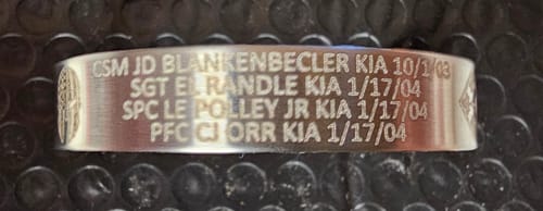 Close-up of a customer's stainless steel bracelet with engraved memorial text for four fallen soldiers, laid on a textured surface.